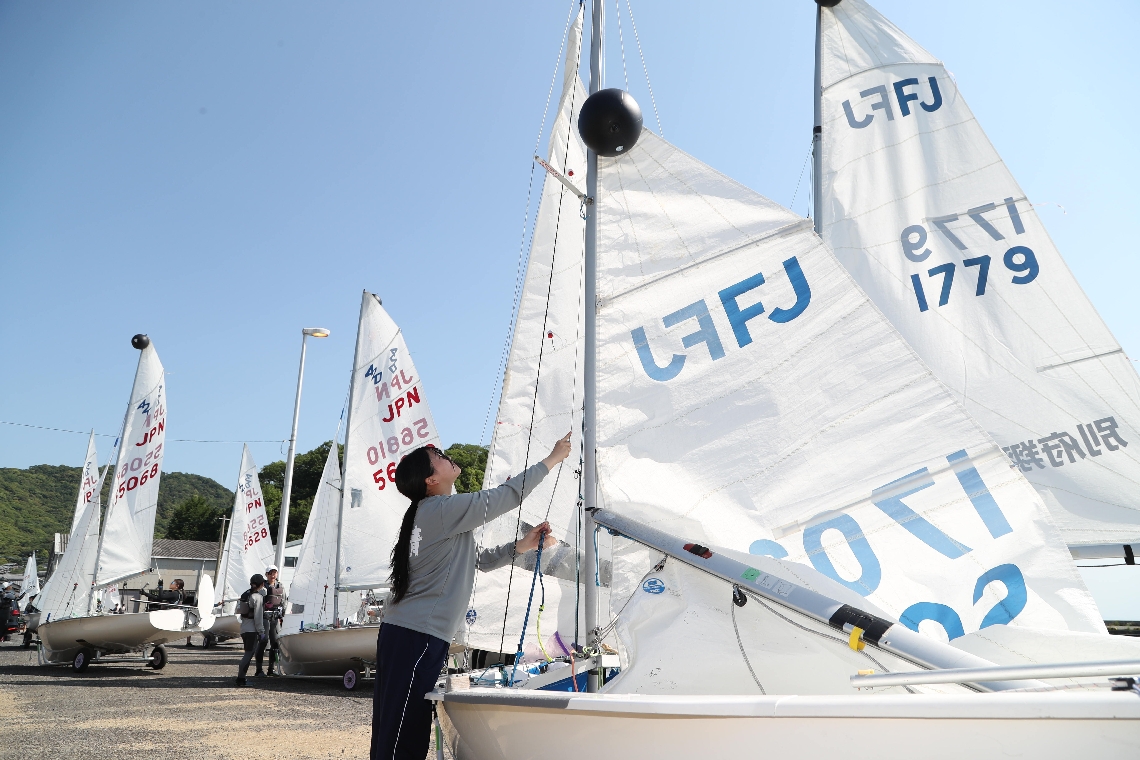 【ヨット女子４２０級】青空の下、海に出る前にヨットの帆を張る選手たち＝臼杵市の海洋科学高艇庫前