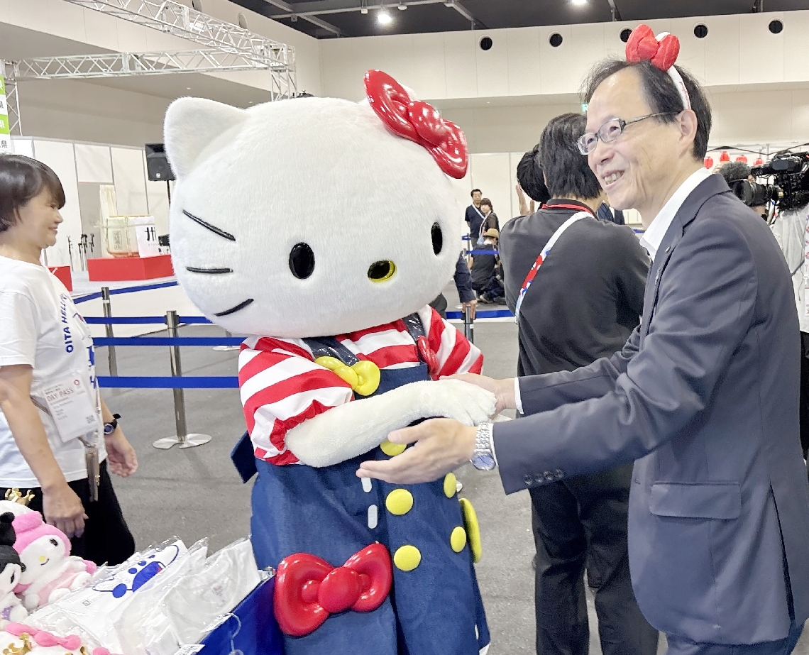 人気キャラクター「ハローキティ」を会場に連れ、日出町のハーモニーランドをＰＲする佐藤樹一郎知事（右）