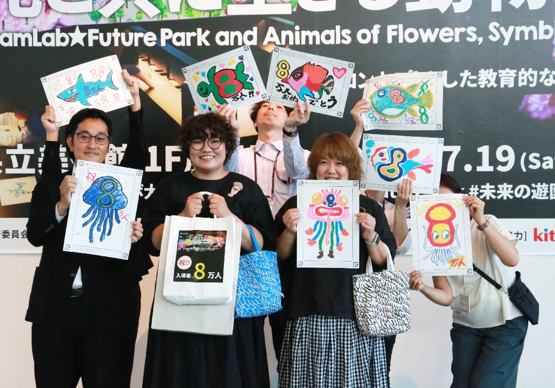 ８万人目になった松尾菜穂さん（前列左から２人目）と母親の智恵さん（同３人目）。県立美術館職員らが祝福のイラストを掲げ祝った＝５日、大分市寿町