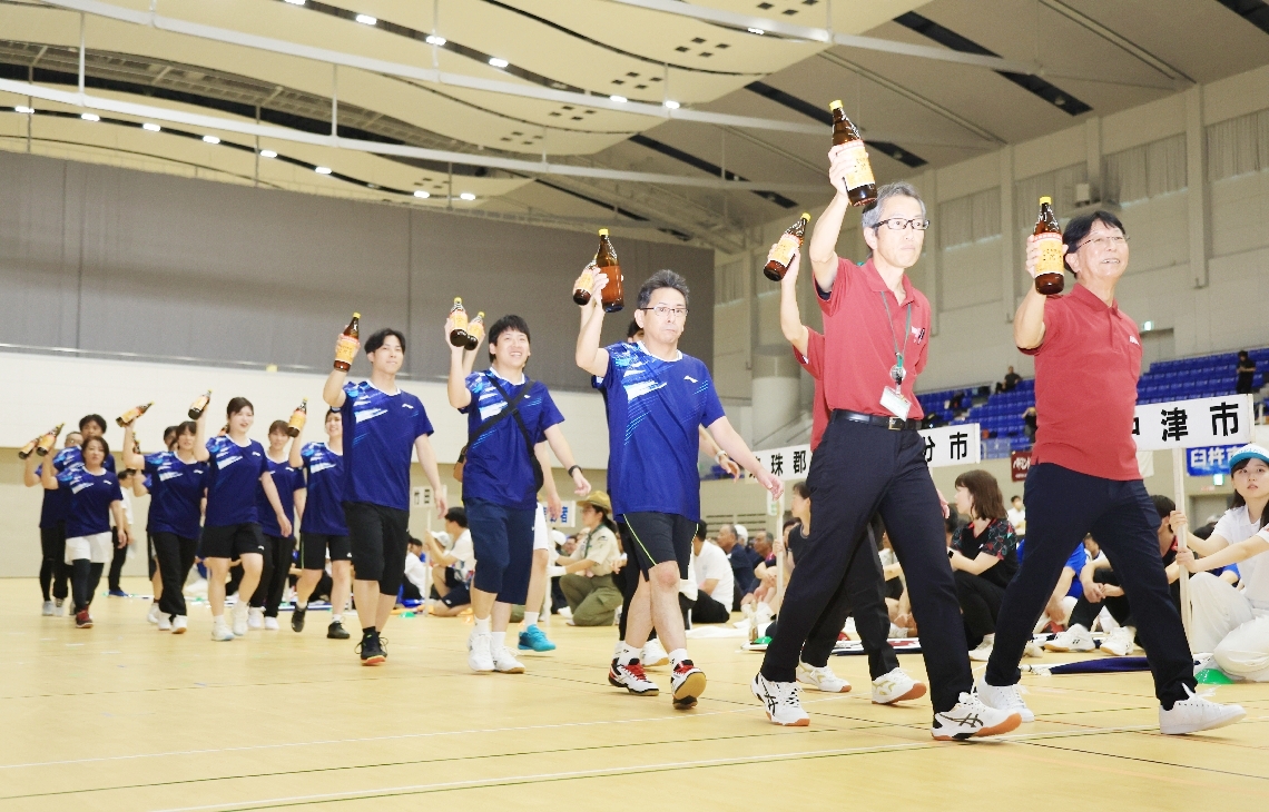 地元の特産品を掲げて入場する選手団＝１３日、べっぷアリーナ
