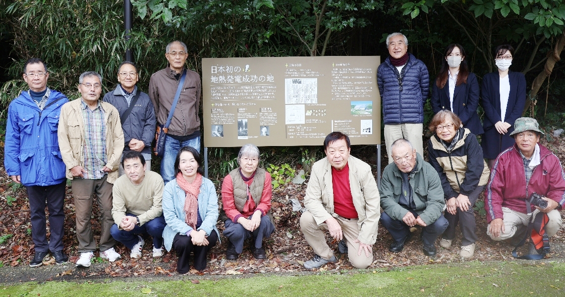 地熱発電の実験成功から１００年の節目に設置した看板。１４人が集まり記念撮影をした＝別府市