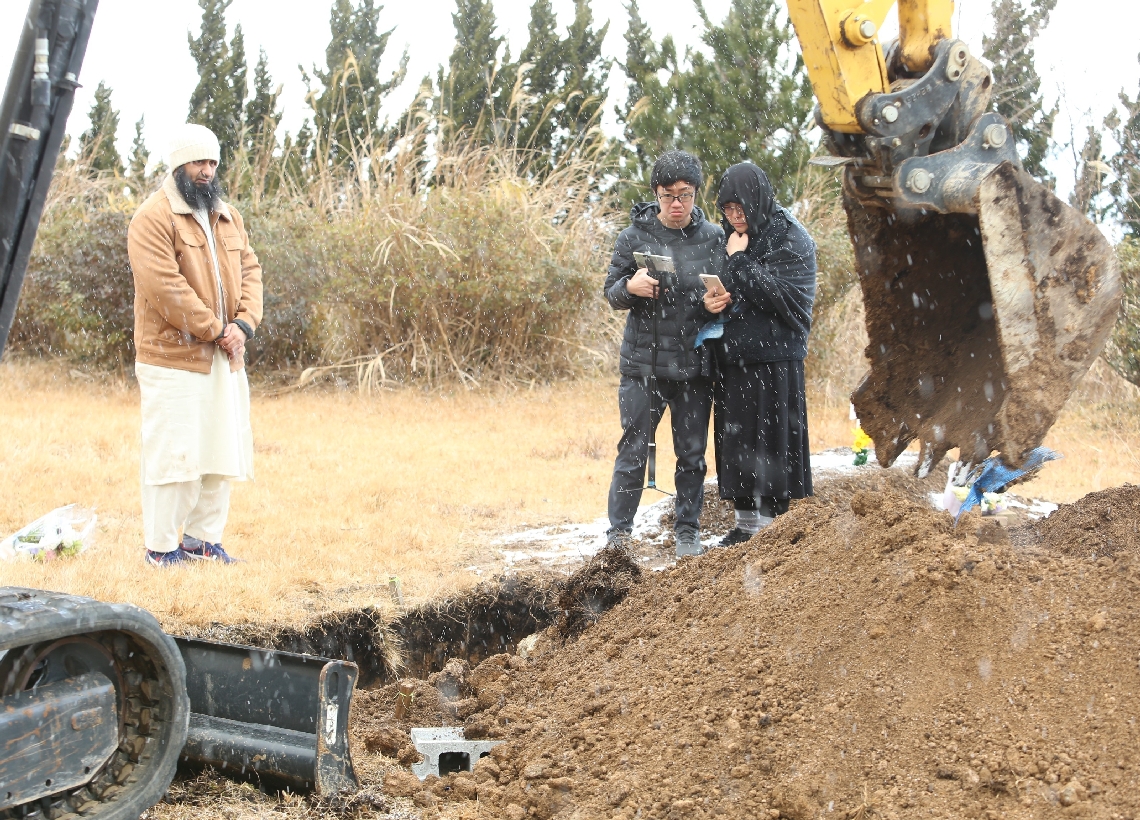 雪が舞う中、ショベルカーがディヤーちゃんの亡きがらに土をかぶせていく。厳しい表情で見守る父親のユハン・クエクさん（左から２人目）と母親の呉超群さん（同３人目）＝１月７日、日出町南畑の大分トラピスト修道院
