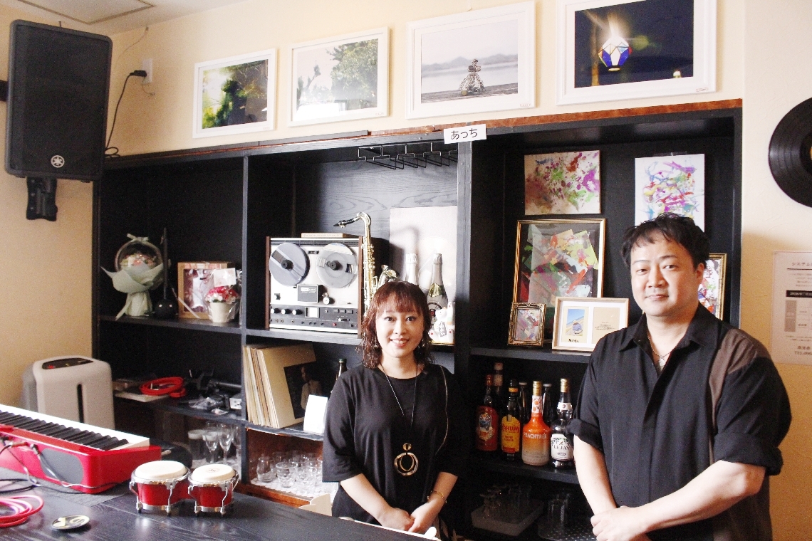 障害のある人による写真や絵画が並ぶスナック。創作活動を支援する翼太郎さん（右）と桜井響子さん＝大分市都町の「Ｏｔｏｄａｎ．」