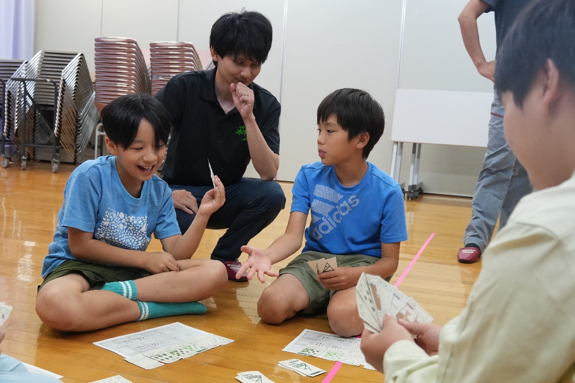 林業のカードゲームに夢中になる児童と、森林インストラクターの都賢太郎さん（左から２人目）