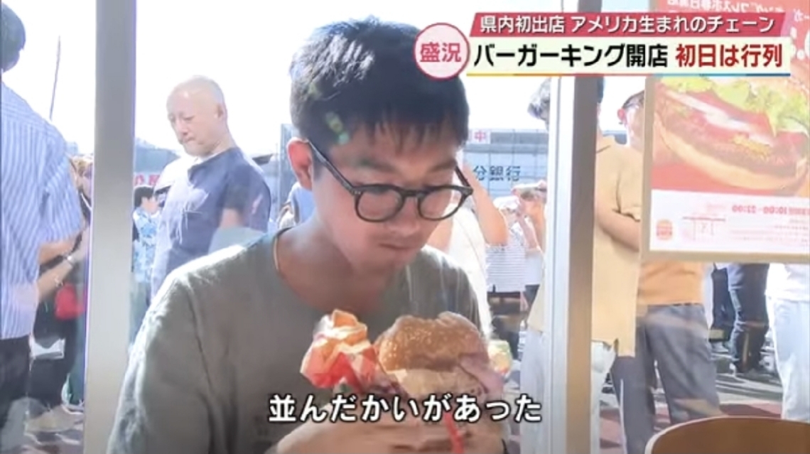 取材に答えながらワッパーを食べる筆者＝大分市（テレビ大分提供）